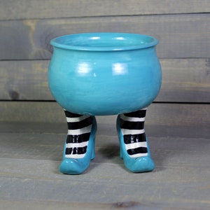 May include: A turquoise ceramic cauldron planter with black and white striped witch legs. The cauldron has a rounded shape and a wide opening. The legs are shaped like high-heeled shoes.