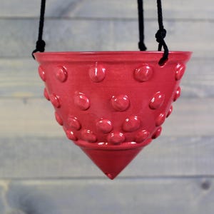 May include: A hanging planter in a vibrant red hue, adorned with raised, rounded embellishments. The ceramic pot has a conical base and is suspended by black rope. This decorative piece is designed for displaying plants.