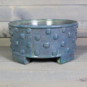 May include: A round ceramic bowl with a glossy blue and teal glaze, featuring a raised polka dot texture across the exterior surface. The bowl is supported by three short, integrated feet at the base, creating a stable and elevated structure.