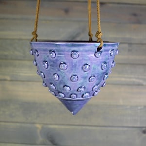 May include: A hanging ceramic planter in shades of blue and purple, adorned with raised, rounded embellishments. The planter is suspended by a natural rope. The design features a cone-shaped bottom and a wide opening, suitable for plants.
