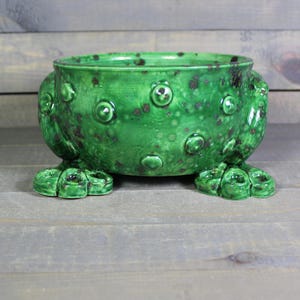 May include: A round, bright green ceramic bowl with a glossy finish and dark brown speckled accents. The exterior features raised, circular bumps and is supported by two wide, textured feet that resemble animal paws on either side.