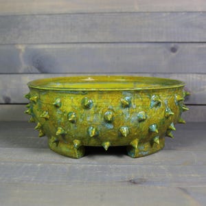 May include: A handcrafted ceramic bowl with a unique, textured design. The bowl is primarily yellow-green with blue-green accents and features numerous cone-shaped spikes around the exterior. It rests on three small, rectangular feet.
