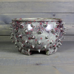 May include: A round ceramic planter featuring a textured surface covered in numerous sharp, protruding spikes. The glaze is a mottled blend of grey, black, and deep red tones, with a smooth interior and a sturdy base with small feet.