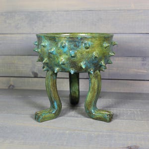May include: A handcrafted ceramic planter with a unique design. The planter is a vibrant green with turquoise accents and features a spiky texture around the rim. It stands on three curved legs, adding to its artistic appeal.