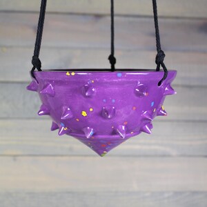 May include: A hanging purple ceramic planter with a spiked exterior and colorful paint splatters. The planter is suspended by black rope and is designed to hold plants. The planter has a unique, modern design.