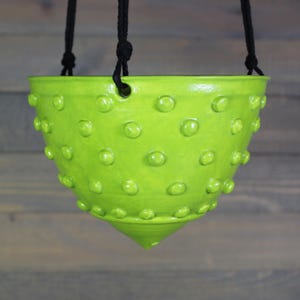 May include: A bright green ceramic hanging planter with a textured, dotted surface. The planter has a conical shape and is suspended by black rope. The vibrant color and unique design make it a decorative piece for indoor or outdoor use.