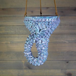 May include: A hanging ceramic planter featuring a textured surface covered in overlapping, iridescent blue and purple petal-like scales. The vessel has a curved, tail-like base and is suspended by three thin brown cords attached to the upper rim.