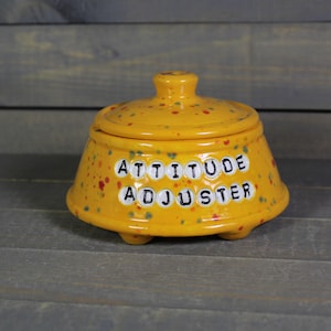 May include: A yellow ceramic jar with a lid, speckled with red, blue, and green paint. The jar has the words "ATTITUDE ADJUSTER" in black letters. The jar sits on three small feet.