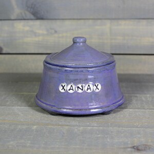 May include: A purple ceramic lidded jar with a glossy finish and a conical lid. The front of the jar features five small white circular beads, each embossed with a black letter, spelling out the word XANAX in a horizontal row across the center.