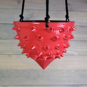 May include: A bright red ceramic hanging planter with a glossy finish and a textured surface covered in numerous small, pointed spikes. The bowl-shaped vessel tapers to a single point at the bottom and is suspended by three black cords.