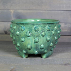 May include: A round ceramic planter with a glossy green glaze and a textured surface featuring numerous raised, rounded bumps. The vessel has a wide opening at the top and is supported by three short, integrated feet at the base.