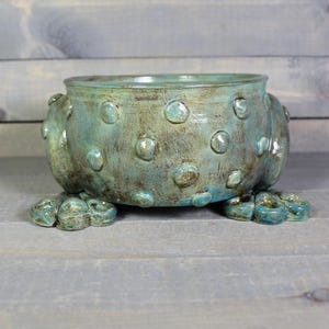 May include: A round ceramic bowl with a teal and brown glazed finish, featuring raised circular dots across the exterior surface. The bowl is supported by three distinct, textured feet that resemble stylized paws or floral shapes.
