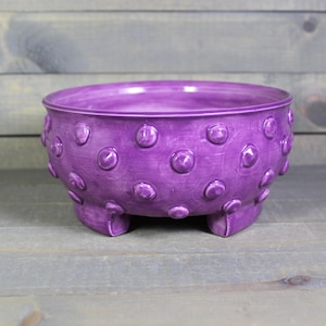 May include: Purple ceramic planter with a textured, dotted surface and three short legs.