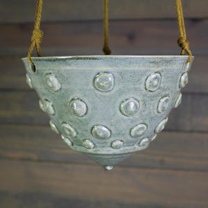 May include: A hanging ceramic planter in a pale blue-green color, adorned with raised, circular patterns. The planter is suspended by a natural rope. The design is a cone shape with a wide opening, suitable for plants.