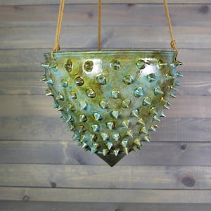 May include: A hanging ceramic planter with a glossy, variegated green and blue glaze finish. The exterior surface is covered in numerous small, sharp, conical spikes, and the bowl is suspended by three thin, light brown braided ropes.
