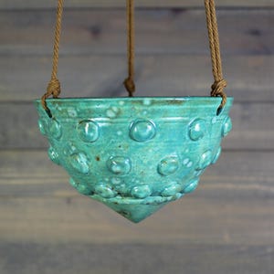 May include: Turquoise ceramic hanging planter with a textured, dotted surface. The planter is suspended by three brown rope cords. The bottom of the planter has a pointed shape. The background is a wooden wall.