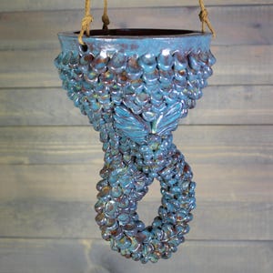 May include: A hanging ceramic planter with a blue and brown glaze. The planter has a textured, scale-like pattern and a butterfly design. It is suspended by a natural rope.
