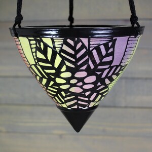 May include: A hanging planter with a black rim and a black, pointed base. The planter's exterior features a design of black leaves and dots on a gradient of yellow and purple. The planter is suspended by a black rope.