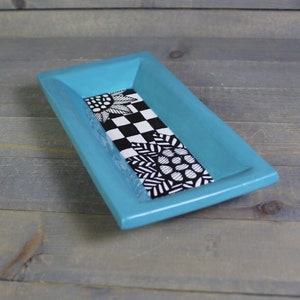 May include: A turquoise rectangular ceramic dish with a black and white patterned design. The pattern features a checkered design, a floral design, and a geometric design.