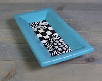 May include: A turquoise rectangular ceramic dish with a black and white patterned design. The pattern features a checkered design, a floral design, and a geometric design.