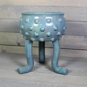 May include: A teal ceramic planter with a rounded bowl featuring raised circular bumps across the exterior surface. The vessel is supported by three long, curved legs that extend downward from the base, finished in a glossy, mottled blue-green glaze.
