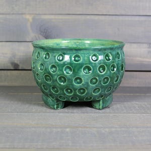 May include: A green ceramic planter with a textured, dimpled surface and three small feet. The planter has a rounded shape and a glossy finish, suitable for small plants or succulents.