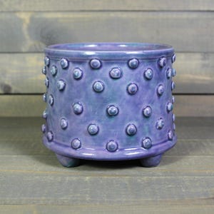 May include: A small, round, ceramic planter in shades of purple and blue. The planter has a textured surface with evenly spaced, raised, circular bumps. It sits on three small feet and is suitable for small plants or succulents.