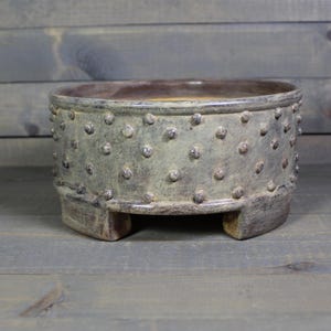 May include: A handcrafted, ceramic planter with a textured surface and a speckled pattern. The planter is a muted green-gray color with a brown interior and sits on three small feet. This decorative piece is suitable for indoor or outdoor use.
