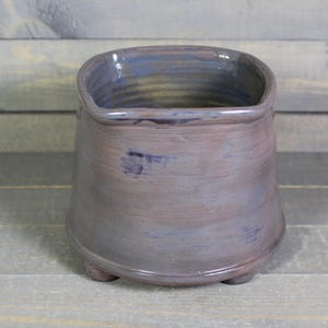 May include: A square-shaped ceramic planter with a light purple glaze and a slightly textured surface. The planter has a wide base and four small feet.