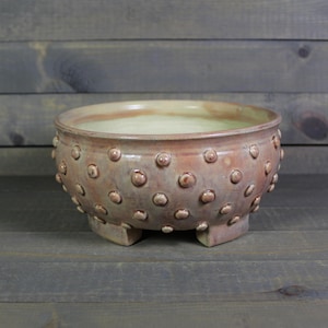 May include: A handcrafted, ceramic bowl in shades of pink and beige, featuring a textured surface with raised, circular accents. The bowl rests on three small, square feet, and is suitable for serving or display.