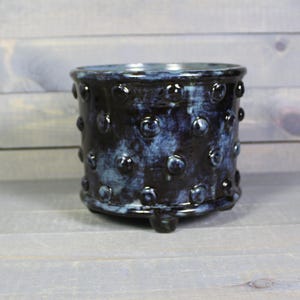 May include: A cylindrical ceramic planter featuring a dark navy blue and black mottled glaze with a glossy finish. The exterior surface is decorated with a repeating pattern of raised, rounded dots and the vessel stands on three small, integrated feet.