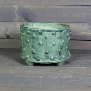 May include: A small, round, green ceramic planter with a textured surface of evenly spaced, raised dots. The planter has three small feet and a slightly flared rim. The color is a muted, earthy green.