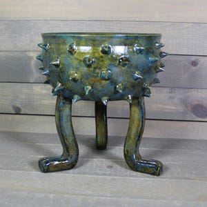May include: A handcrafted ceramic planter with a unique design. The blue and green glazed pot features a textured surface with protruding spikes. It stands on three curved legs, adding a whimsical touch to the decorative piece.
