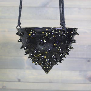 May include: A black ceramic hanging planter with a conical shape and a glossy finish. The surface is covered in numerous sharp, protruding spikes and features a speckled pattern of small gold-colored paint flecks. It hangs from black cords.