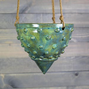 May include: A hanging ceramic planter with a textured surface and a cone-shaped bottom. The planter is glazed in shades of green and blue, with raised circular details. It hangs from a natural rope.