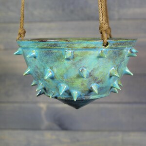 May include: A bowl-shaped ceramic hanging planter with a glossy turquoise and green glaze. The exterior surface is covered in numerous pointed, cone-shaped spikes. The planter is suspended by two thick, brown, knotted jute ropes attached to the rim.