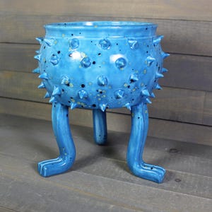 May include: A bright blue ceramic planter with a rounded body covered in numerous small, pointed spikes. The vessel features a speckled finish with dark spots and is supported by three long, curved legs that extend outward from the base.