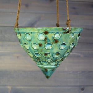 May include: A cone-shaped ceramic hanging planter with a glossy teal and green glaze. The exterior surface features a repeating pattern of circular indentations and dark brown speckles. It is suspended by three thick, natural-colored twisted ropes.