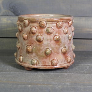 May include: A handcrafted ceramic planter with a textured surface of raised, rounded bumps. The planter is a warm, earthy color with hints of green and brown. It has a cylindrical shape and sits on three small feet. A decorative piece for plants.