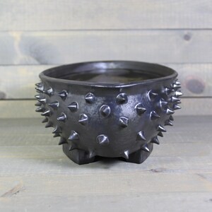 May include: A matte black ceramic bowl featuring a wide, open top and a rounded body covered in numerous sharp, protruding spikes. The bowl is supported by three small, sturdy feet at the base, creating a distinct, textured appearance.