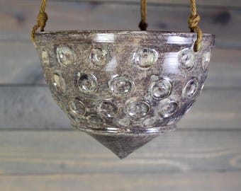 May include: A hanging ceramic planter with a textured, mottled finish in shades of gray and green. The planter features a series of raised, circular designs and is suspended by three woven rope cords. The planter has a conical base.
