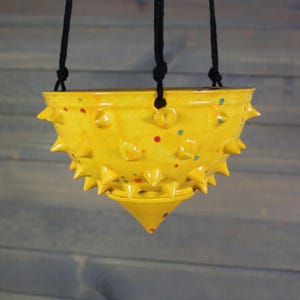 May include: A hanging yellow ceramic planter with a spiky design. The planter has a conical bottom and is suspended by black rope. It is decorated with red and blue speckles.