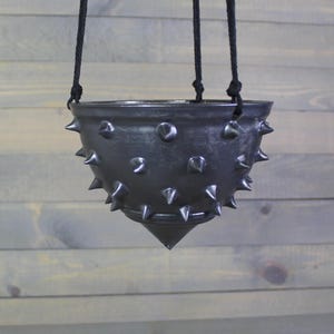 May include: A dark gray, hanging planter with a spiked exterior. The ceramic pot is suspended by three black cords. The planter has a conical bottom and is suitable for small plants or decorative purposes.