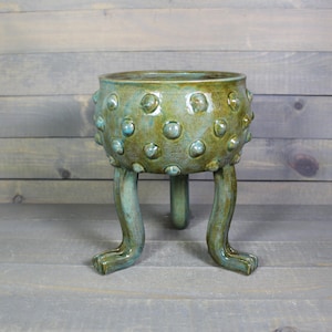 May include: A ceramic planter with a glossy, mottled green and blue glaze. The bowl features raised, rounded bumps across its exterior surface and is supported by three curved, leg-like appendages that extend downward from the base of the vessel.