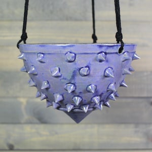 May include: A bowl-shaped ceramic hanging planter with a glossy purple glaze and numerous protruding spikes covering the exterior surface. The planter is suspended by black cords attached to holes along the upper rim of the vessel.