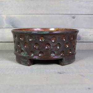 May include: A handcrafted ceramic bowl with a dark brown glaze and a pattern of evenly spaced, circular indentations. The bowl has a slightly flared rim and sits on a low, rectangular base. The color transitions to green and yellow near the rim.