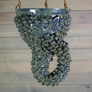 May include: A hanging ceramic planter with a blue-green glaze. The planter is adorned with a textured, bubble-like pattern and a leaf design. It is suspended by a brown rope.