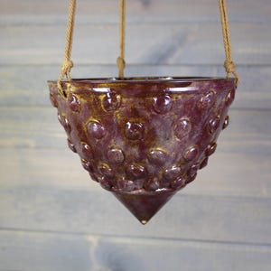 May include: A conical ceramic hanging planter with a deep burgundy glaze and gold-toned highlights. The exterior surface features a repeating pattern of raised circular bumps. The planter is suspended by three tan-colored twisted fiber ropes.