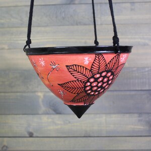 May include: A conical hanging planter with a glossy black rim and pointed base. The exterior is a vibrant coral orange featuring a large, hand-painted black sunflower motif with white dots in the center, surrounded by small white dandelion seed patterns.
