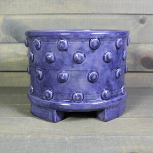 May include: A purple ceramic planter with a textured, dotted design and four small feet.